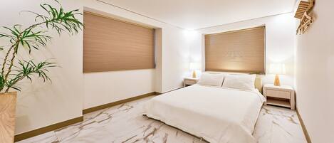 A modern bedroom with a large bed, two bedside tables, a nightstand, a potted plant, and large windows with blinds.