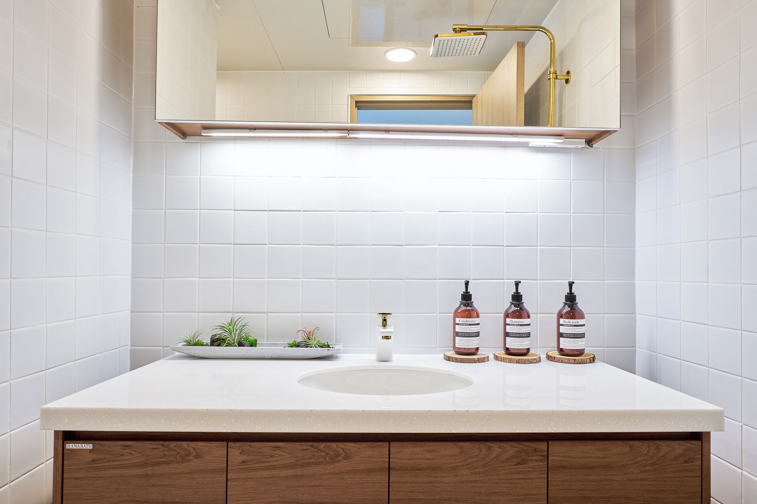 A bathroom with white tiles, a sink, a mirror, and a shower. There are three bottles of soap on the counter.
