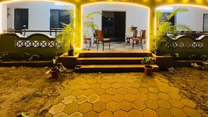 Entrada de un hotel con un camino de baldosas hexagonales que lleva a un conjunto de escalones. Hay plantas en macetas a ambos lados del camino, y se ve un área de descanso con sillas y mesas al fondo.