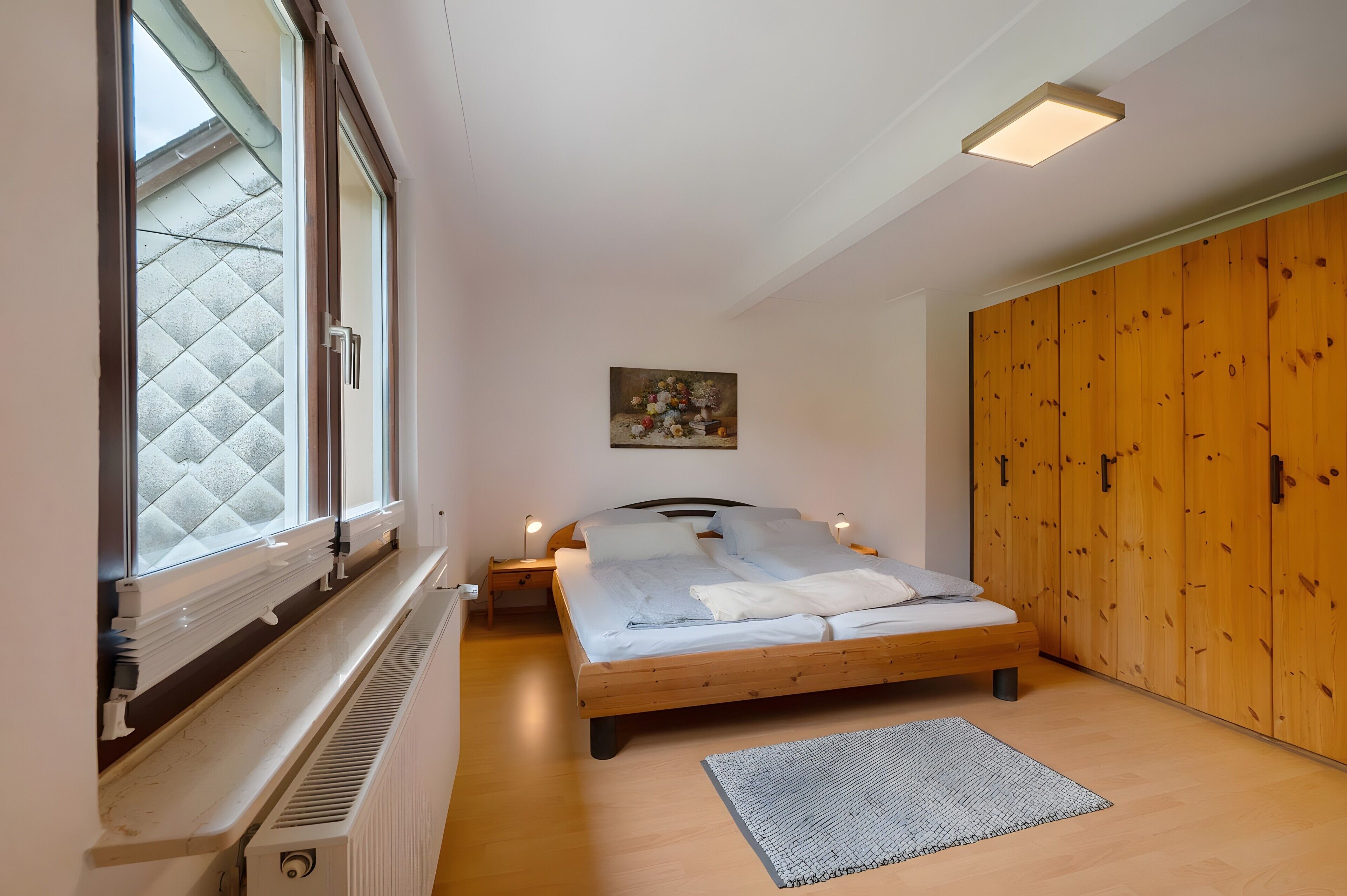 A bedroom with a bed, a radiator, a window, a painting, and wooden wardrobe doors.