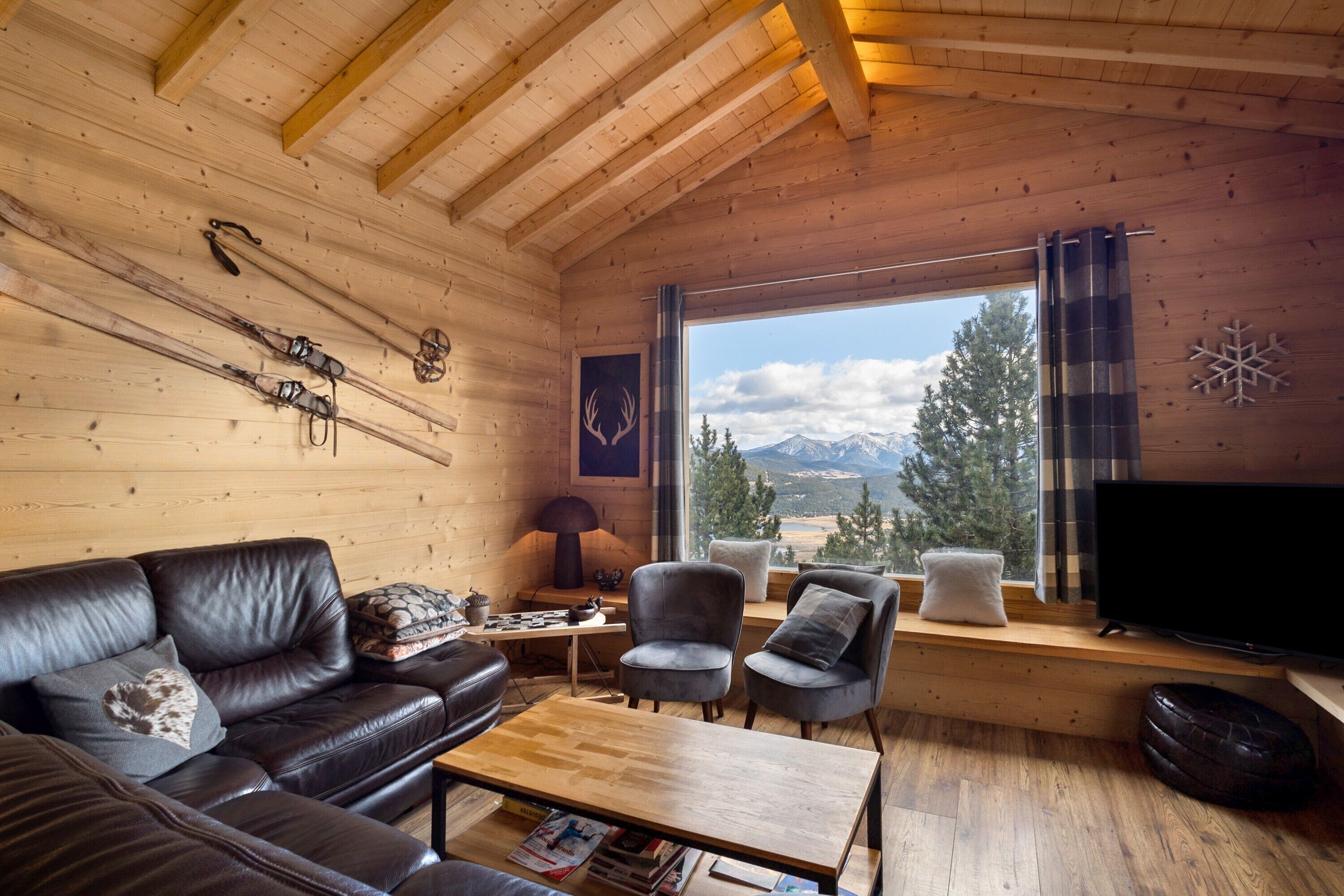 A wooden cabin interior with a sofa, armchairs, a table, skis, a TV, and a window with a mountain view.