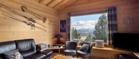 A wooden cabin interior with a sofa, armchairs, a table, skis, a TV, and a window with a mountain view.