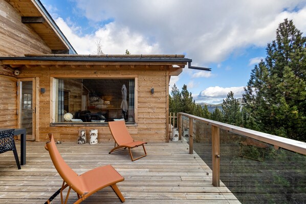 A wooden cabin with a deck, two lounge chairs, and a glass railing overlooking a forested area.
