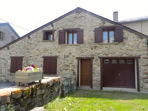 Un bâtiment en pierre avec une porte marron, deux fenêtres avec volets et un petit parterre de fleurs.
