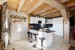 Une cuisine avec des poutres en bois, des meubles blancs, un îlot central avec des tabourets et des ustensiles de cuisine suspendus.