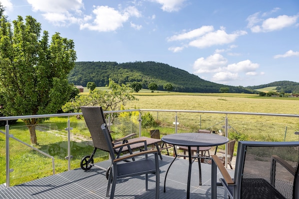A terrace with outdoor furniture, a glass railing, and a view of a green field and hills. - Apartment 'Lerchenweg B' with Shared Terrace, Shared Garden and Wi-Fi (Bad Wildungen)