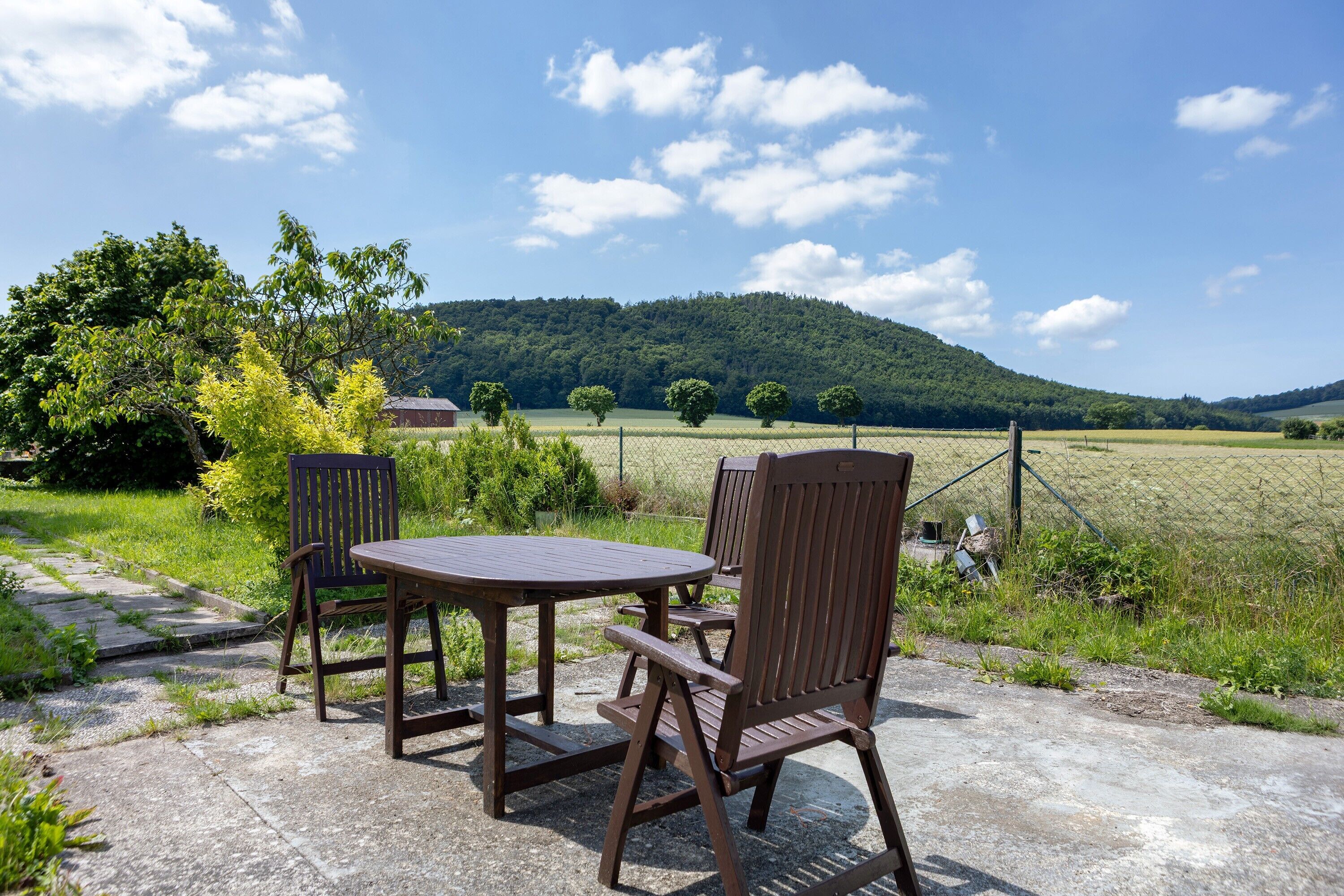 En udendørs terrasse med et rundt bord og to stole, omgivet af græs og et hegn.