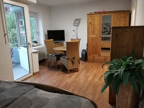 A room with a wooden floor, a dining table with chairs, a television, a cabinet, and a large window.