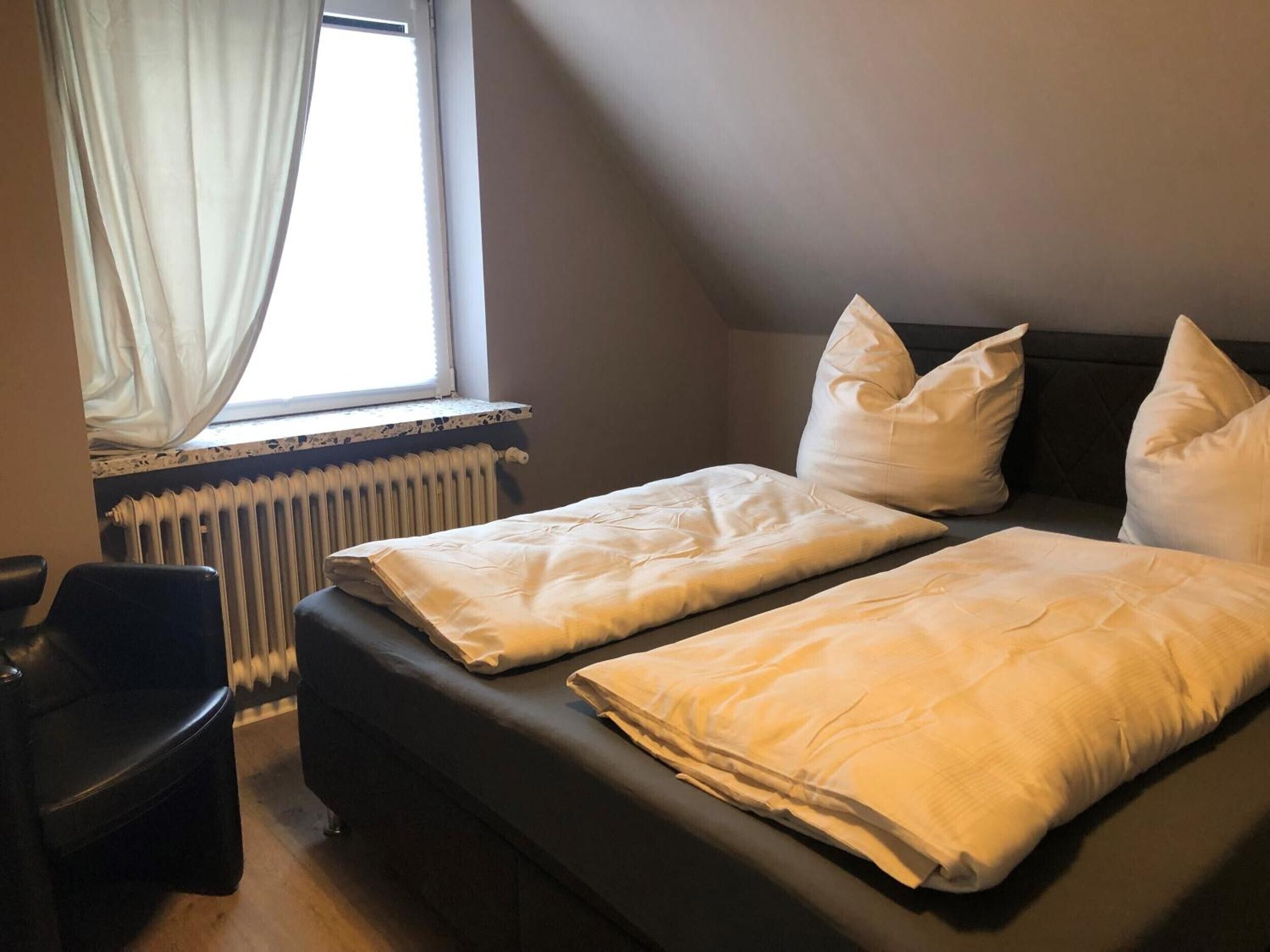 A bedroom with a bed, pillows, a chair, a window with curtains, and a radiator.