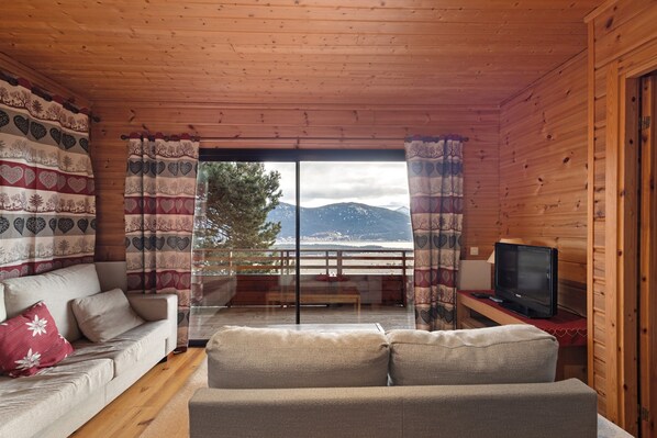 Interior de una cabaña de madera con sofá, televisor y vista al océano.