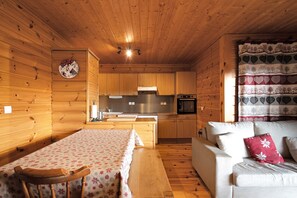 A wooden cabin interior with a dining table, a sofa, a kitchen area, and a decorative wall clock.