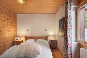 A bedroom with a wooden ceiling, a bed with white bedding, two bedside lamps, a window with curtains, and a wall-mounted picture. - Chalet le Cerf - 3 Bedrooms, South-Facing Terrace with Stunning View, 1 km from Slopes (Les Angles)