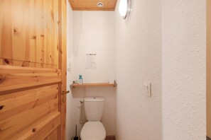 Un baño pequeño con puerta de madera, un inodoro, un estante con artículos de aseo y una pared blanca.