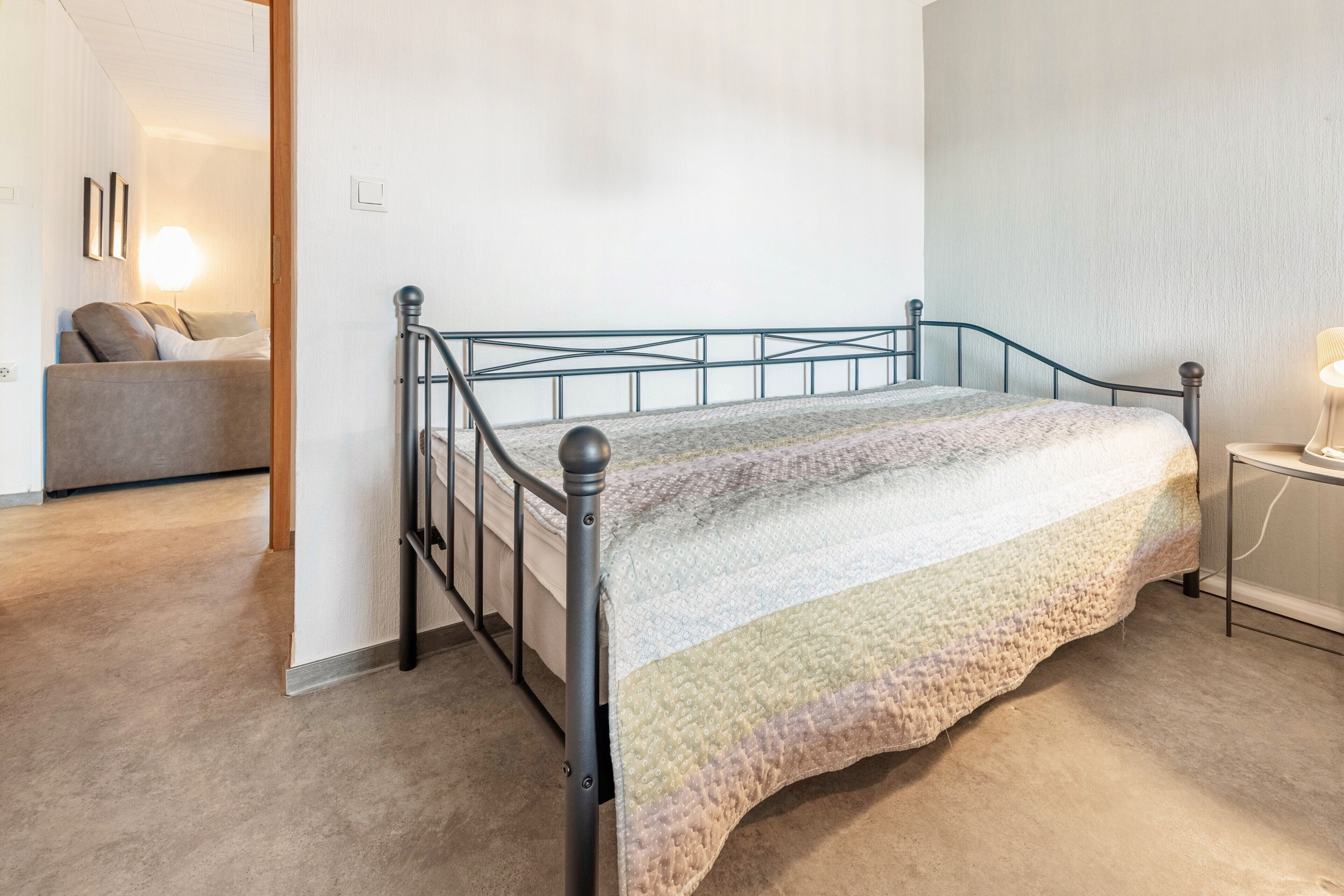 A bedroom with a metal bed frame, a quilted bedspread, a bedside table with a lamp, and a sofa in the adjacent room.