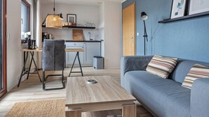 A modern living room with a grey sofa, a wooden coffee table, a grey chair, a kitchen area with a hanging light fixture, and a blue accent wall. - Apartment 'Hochwald' with Shared Terrace, Shared Garden and Wi-Fi (Bad Sachsa)