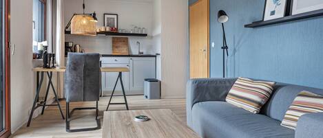 A modern living room with a grey sofa, a wooden coffee table, a grey chair, a kitchen area with a hanging light fixture, and a blue accent wall.