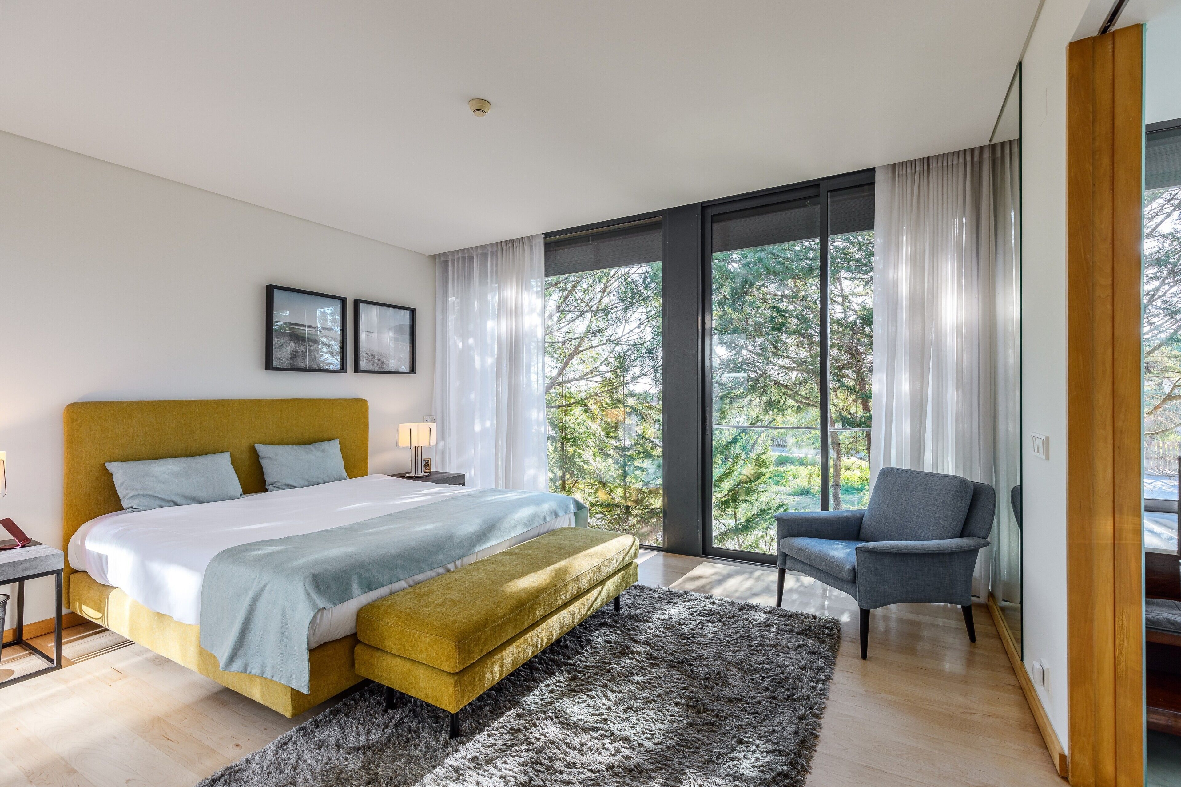 Een moderne slaapkamer met een bed, een bankje, een stoel, een nachtkastje en een groot raam met gordijnen.