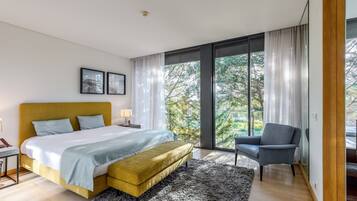 Een moderne slaapkamer met een bed, een bankje, een stoel, een nachtkastje en een groot raam met gordijnen.