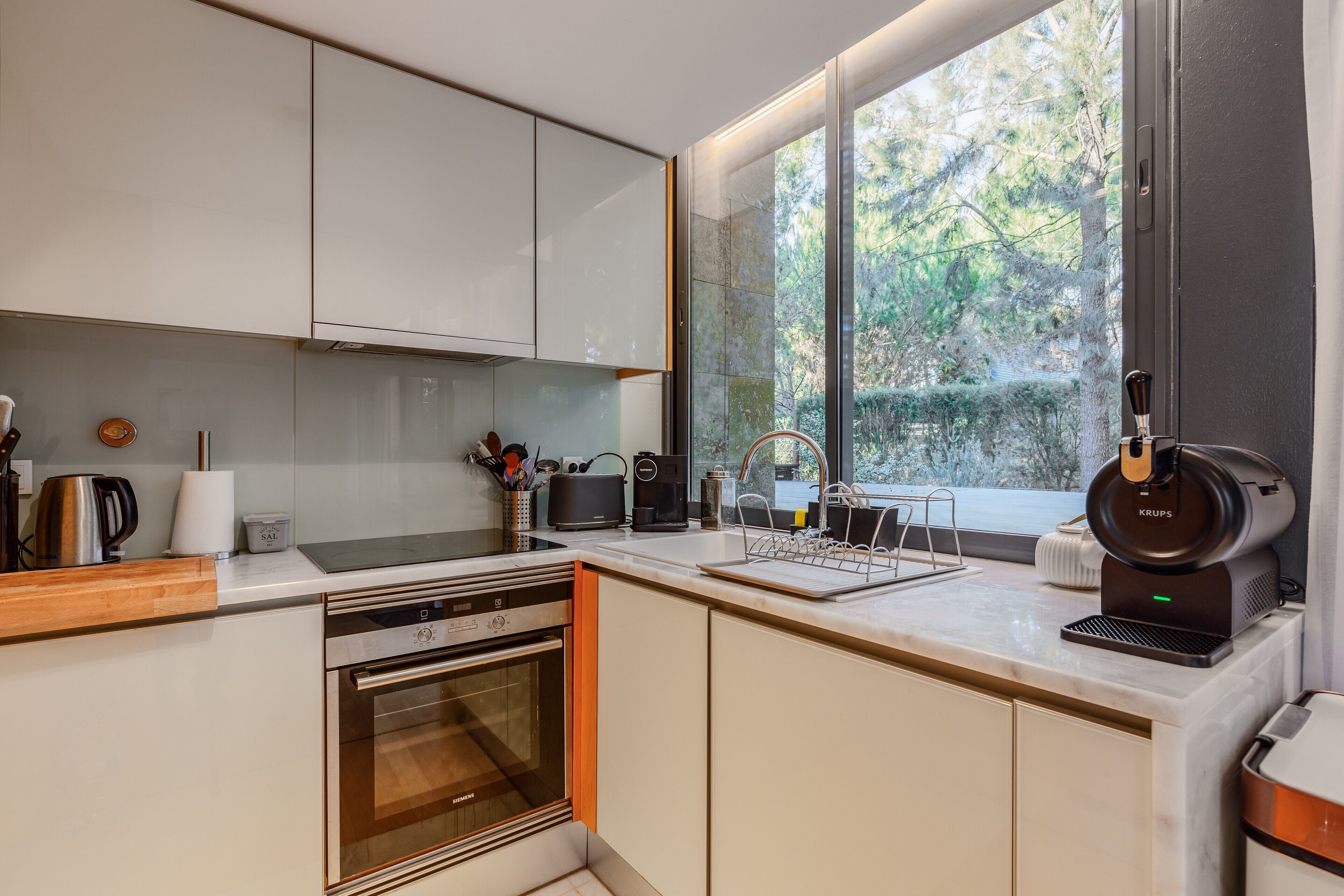 A modern kitchen with a built-in oven, a coffee machine, and a window with a view of trees.