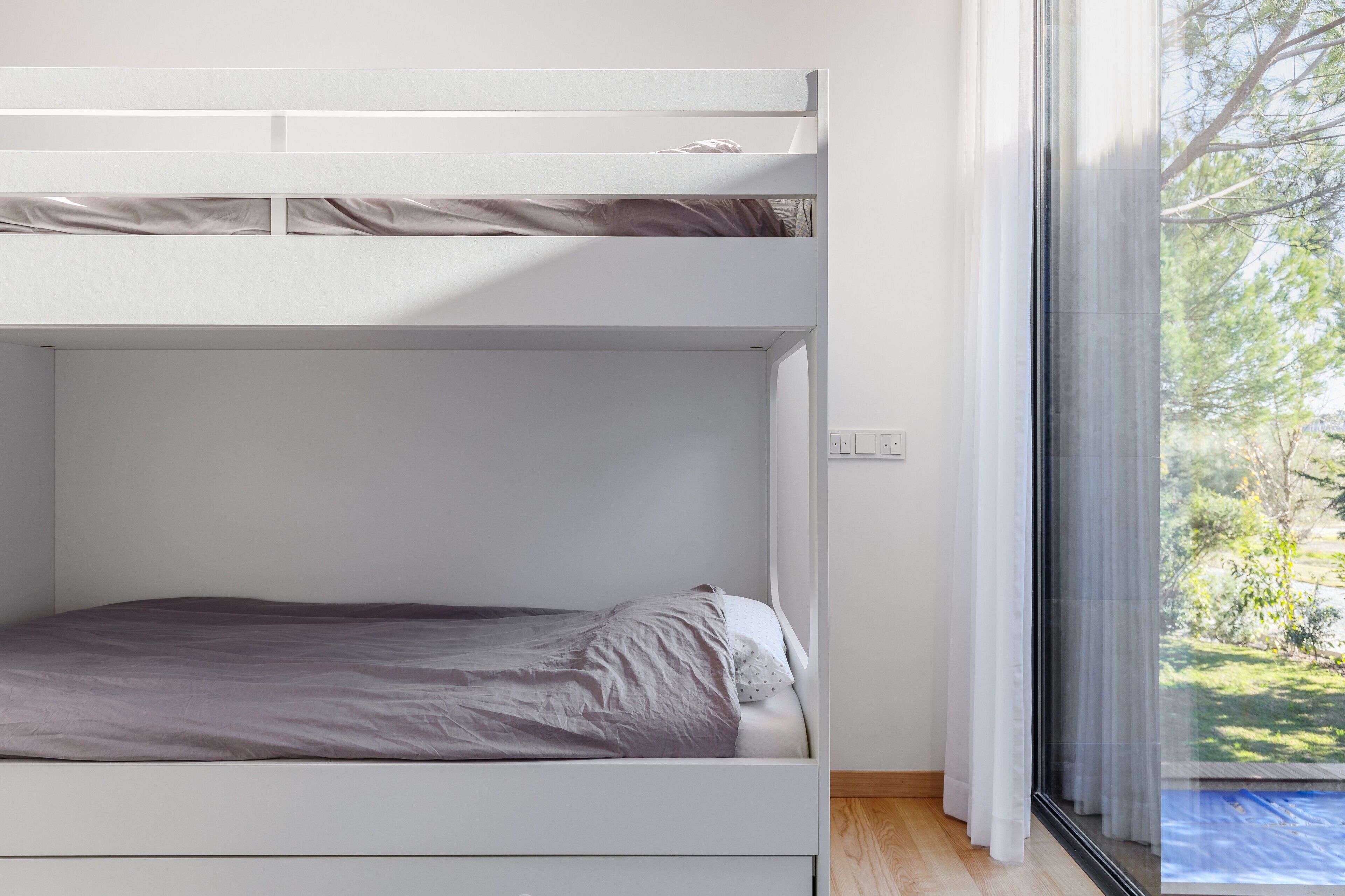A bunk bed with a window, a wooden floor, and a view of greenery outside.