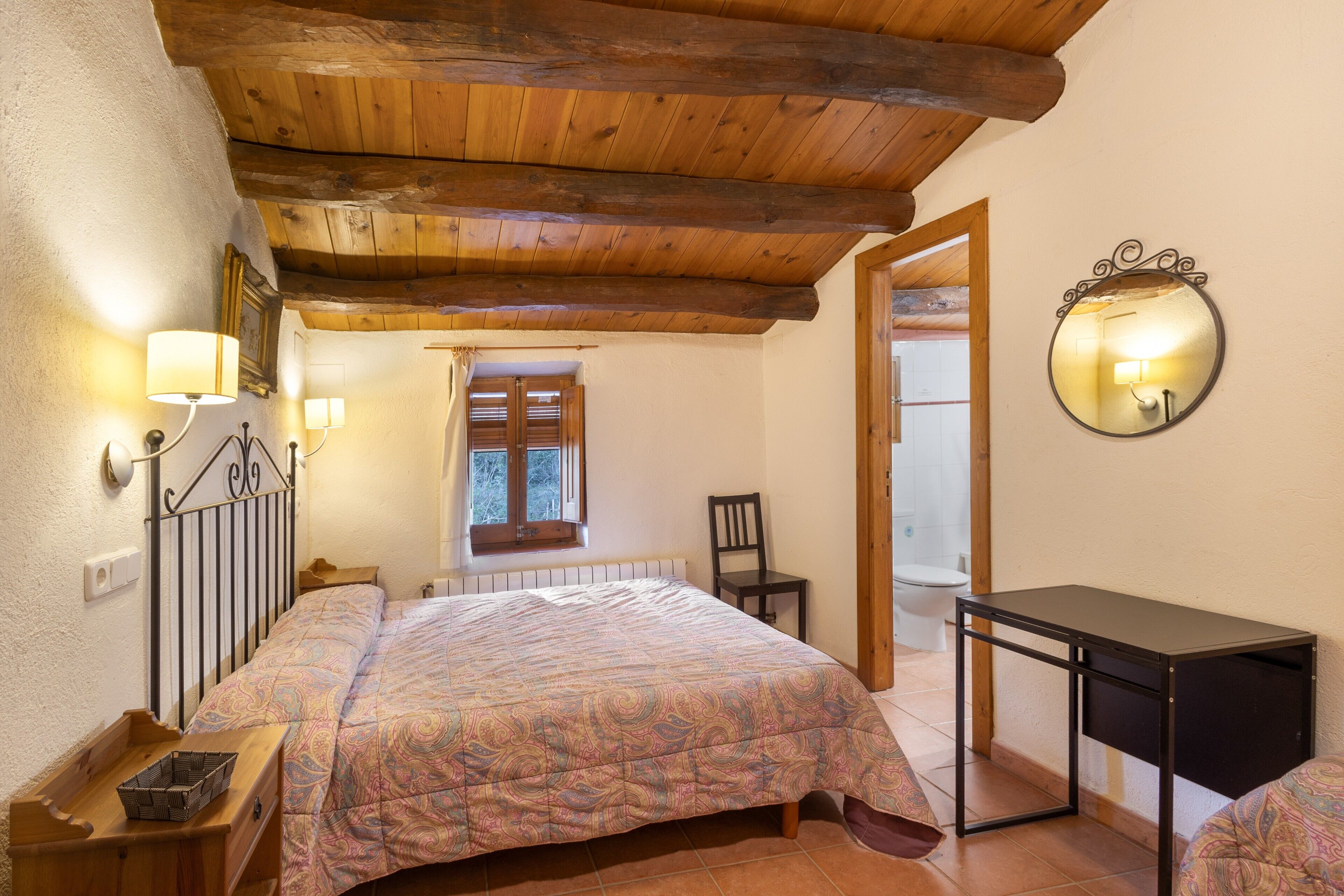 Un dormitorio con techo de madera, una cama con colcha estampada, una mesita de noche, una silla, un escritorio y un espejo en la pared.