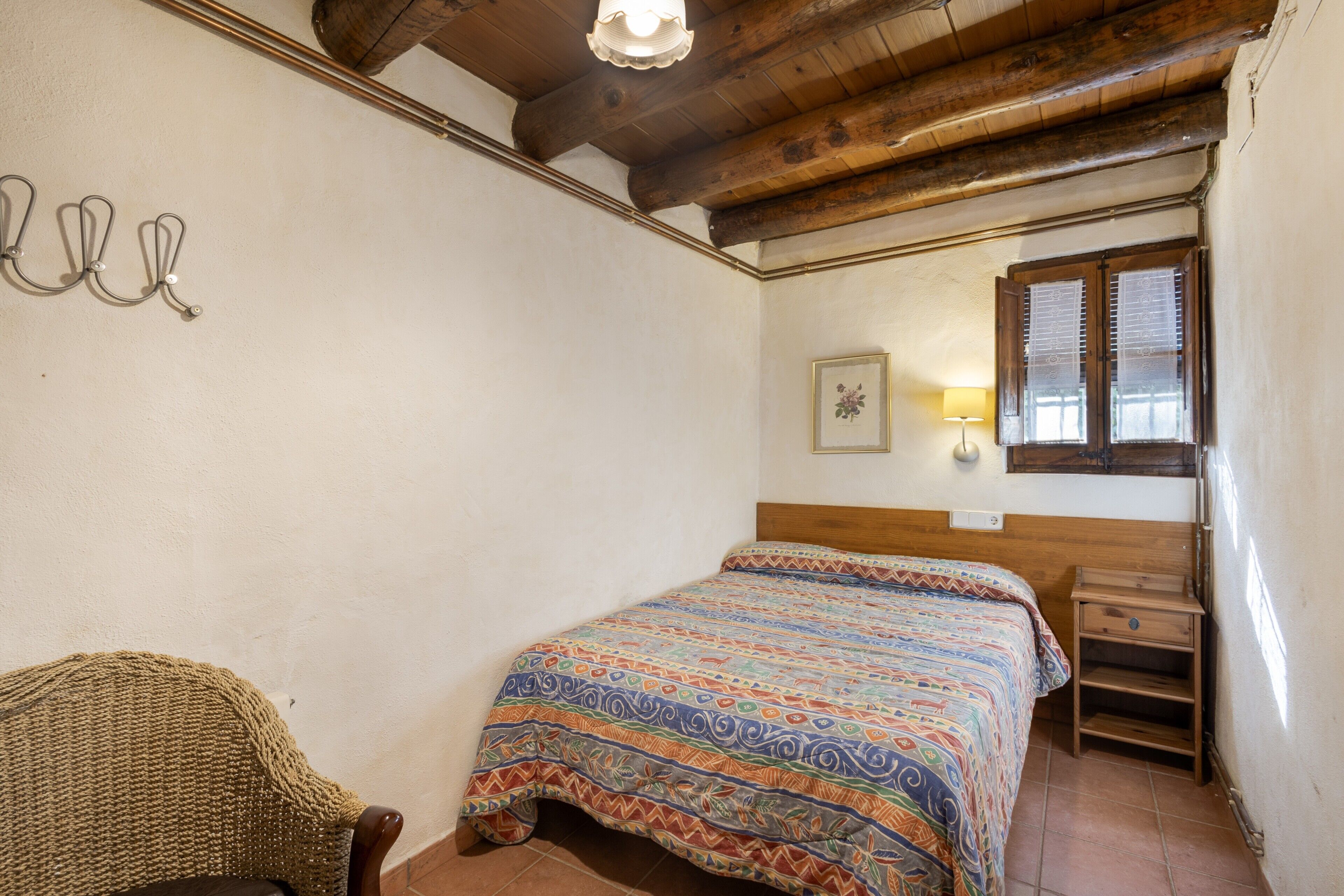 A bedroom with a wooden ceiling, a bed with a patterned bedspread, a chair, a nightstand, and a window with blinds.