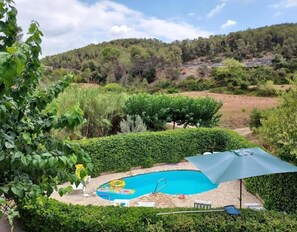 Une piscine dotée d’un parasol, entourée de verdure et avec des collines en arrière-plan.