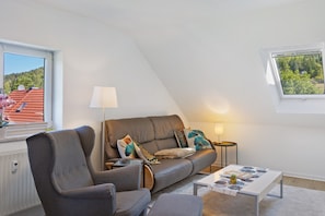 Ett modernt vardagsrum med en soffa, fåtölj och soffbord, en lampa och utsikt över en byggnad med rött tak genom ett fönster.