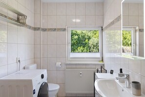 Une salle de bain avec une fenêtre, un radiateur, des toilettes, un lave-linge et un lavabo.