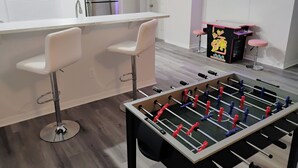 A modern kitchen with a foosball table, two bar stools, a refrigerator, and a neon sign.