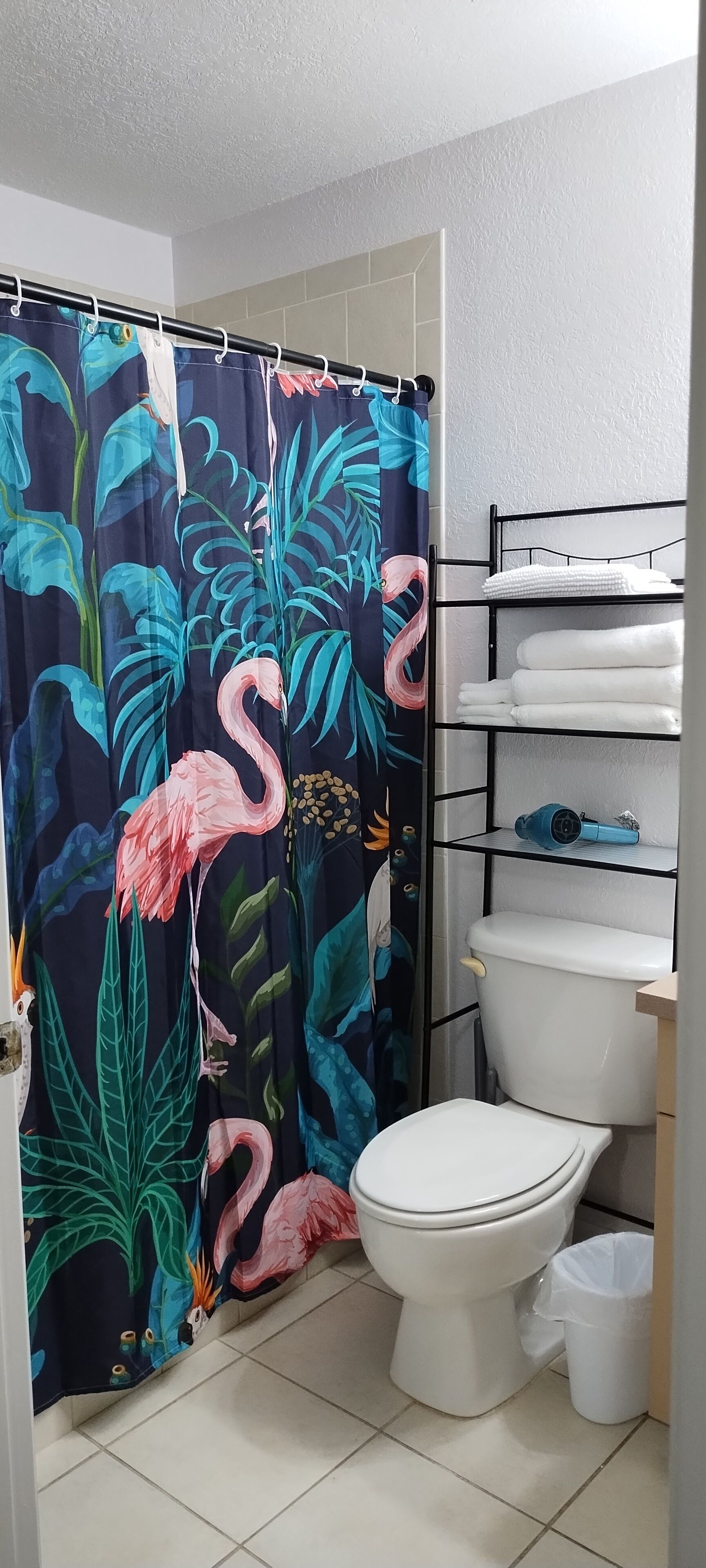 A bathroom with a toilet, a shelf with towels, and a shower curtain with a tropical design.