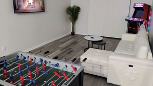 A living room with a foosball table, a white sofa, a small round table, and a wall-mounted TV displaying a fireplace scene.