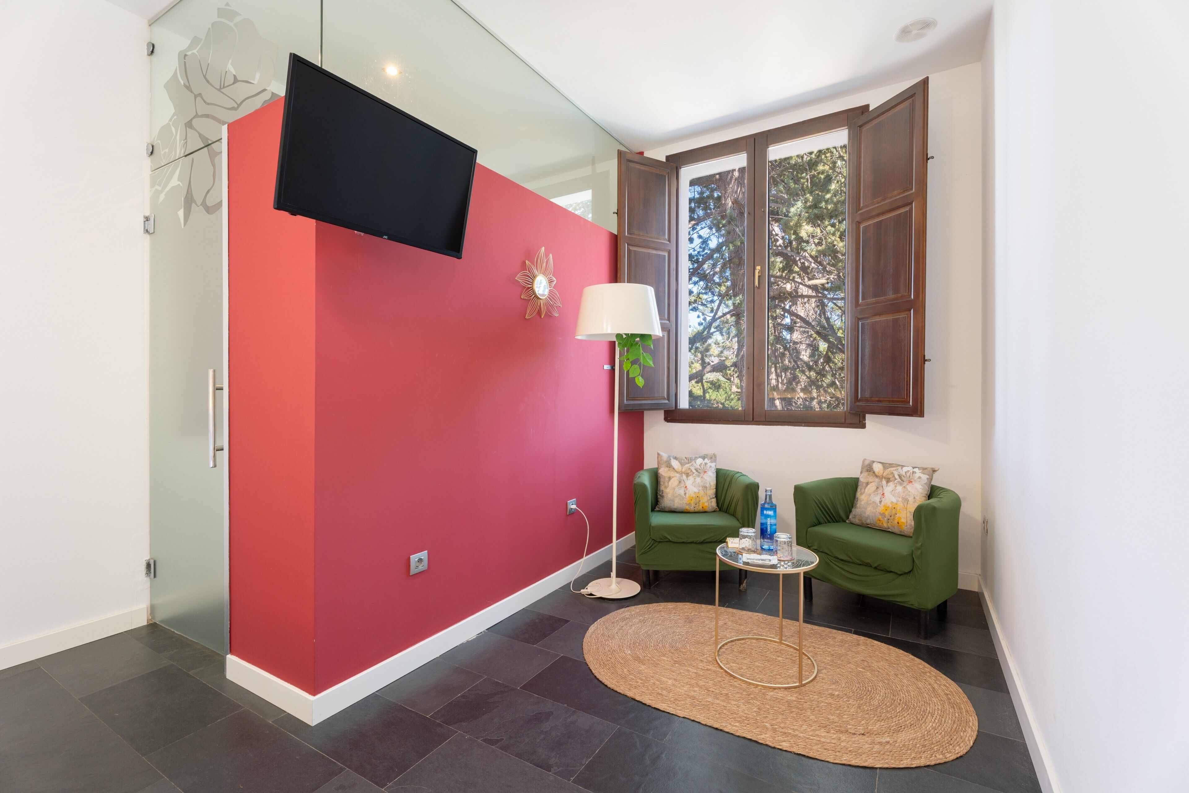 Una habitación moderna con una pared de acento roja, un televisor de pantalla plana, un sillón verde, una mesita redonda y un ventanal con vistas a árboles.
