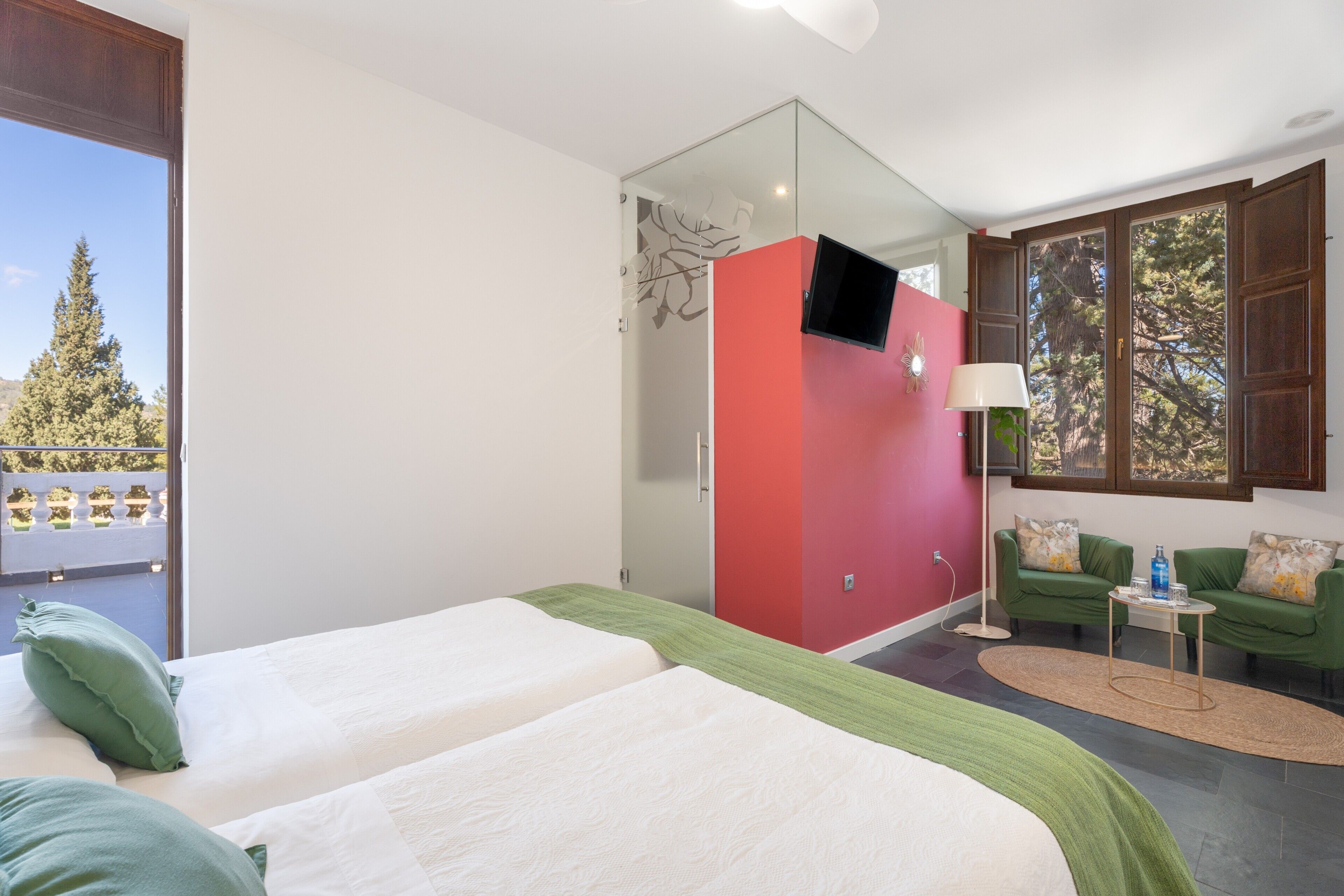 Une chambre d’hôtel avec deux lits, une armoire rouge, une télévision, un canapé vert et une vue sur les arbres.