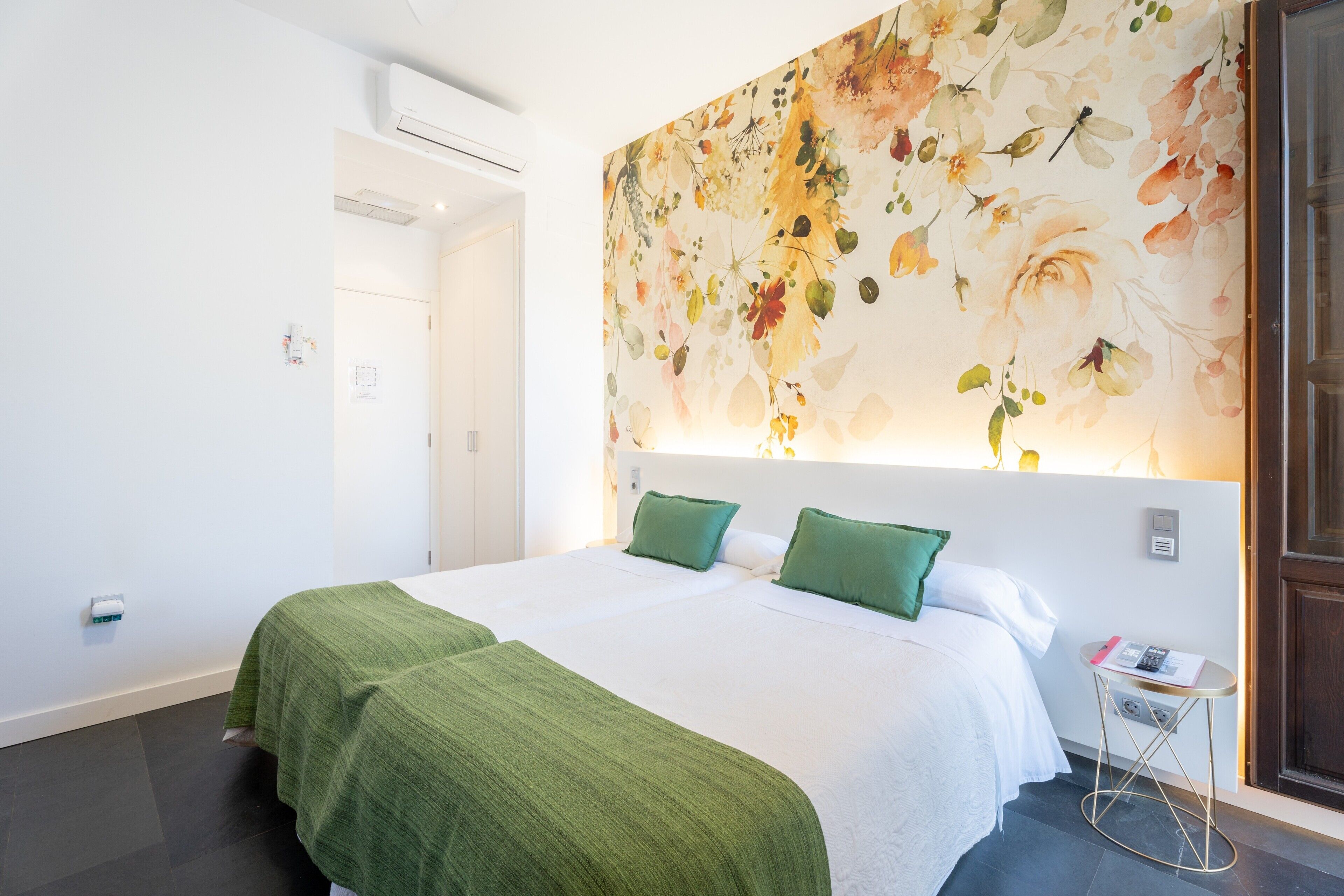 A hotel room with a large floral mural, a bed with green bedding, a bedside table with a phone, and a door.