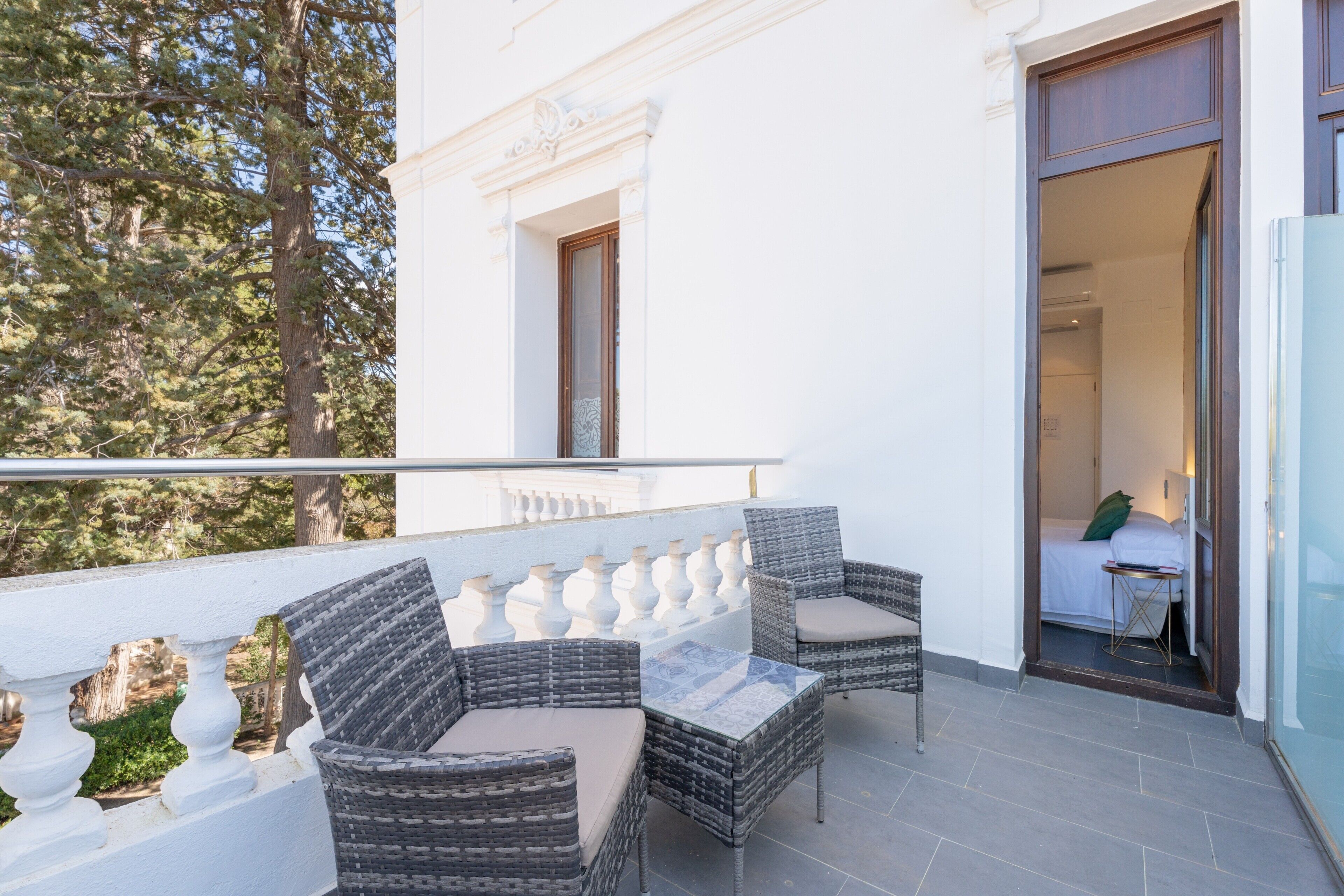 A balcony with wicker furniture, a glass table, and a view of trees.