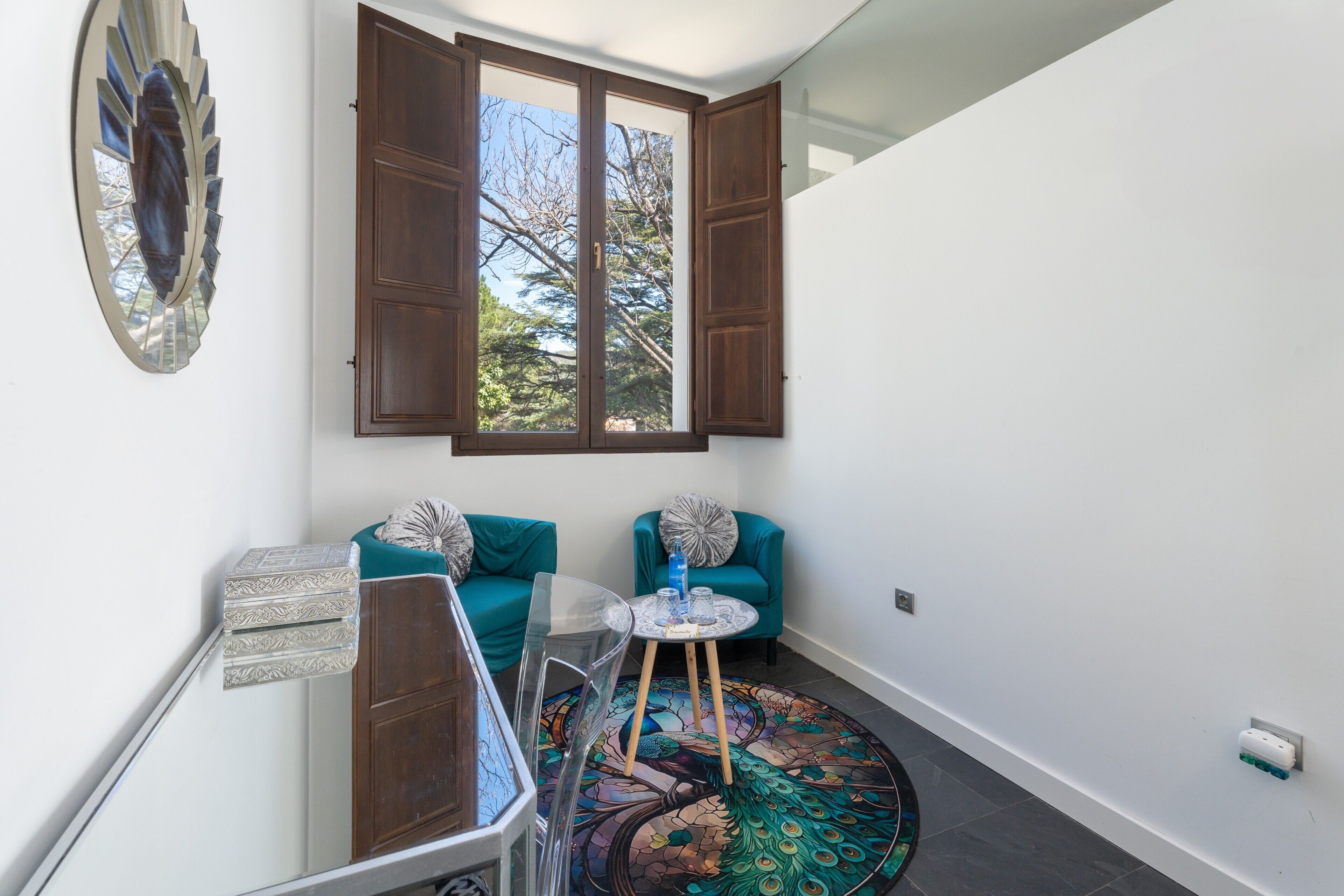 Una habitación con un ventanal grande, sillas color turquesa, una mesita redonda y una alfombra con motivos de pavo real.