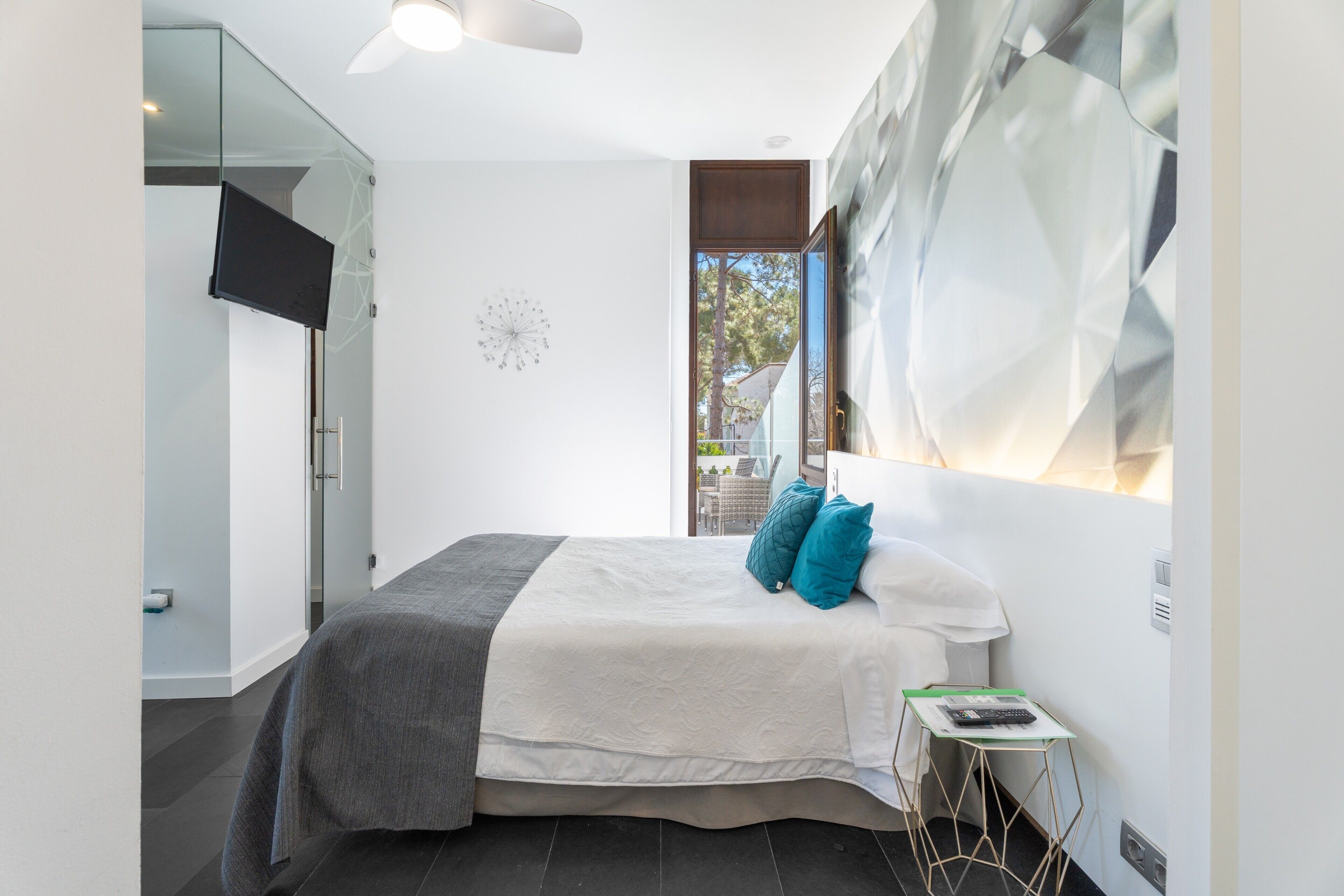 Un dormitorio moderno con cama, televisor, puerta de cristal que da a un balcón y un diseño geométrico en la pared.