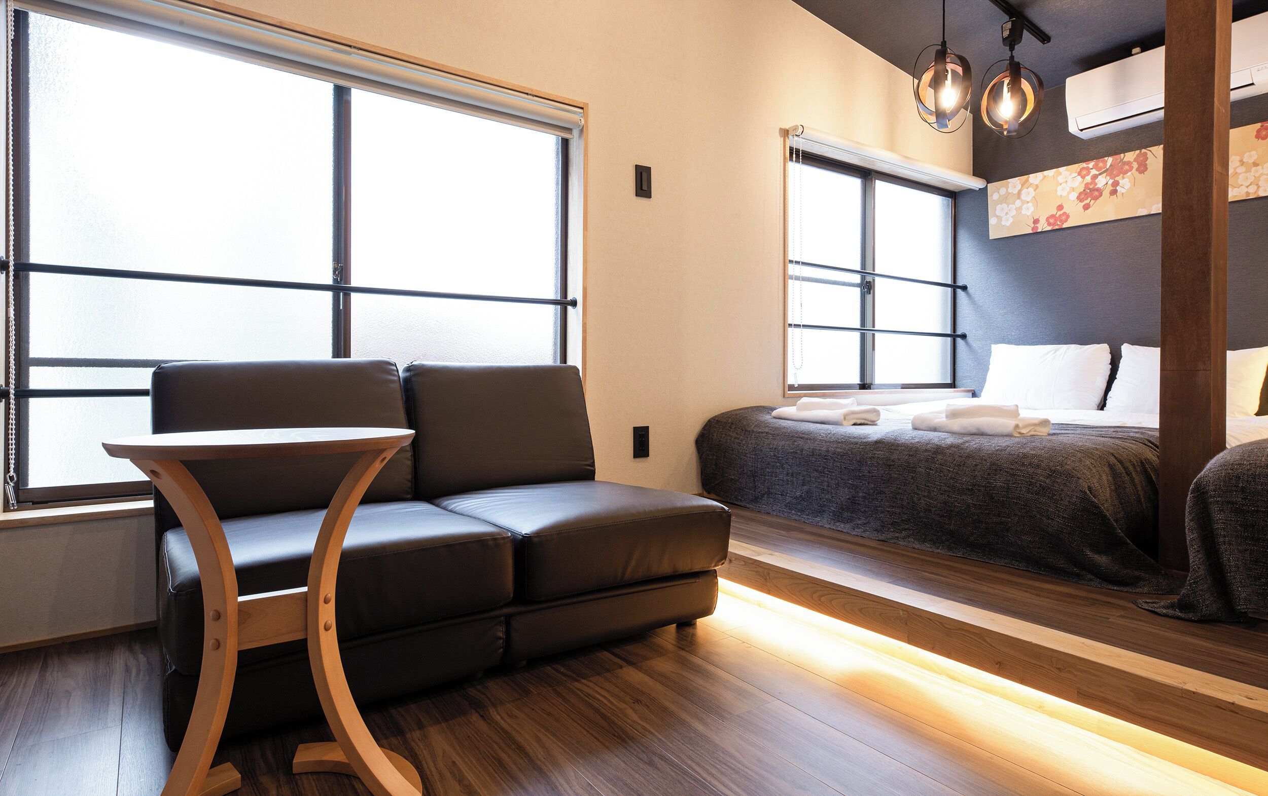 A modern hotel room with a sofa, a small table, a bed with neatly made linens, and large windows providing natural light.
