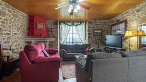 A cozy living room with stone walls, a red armchair, a grey sofa, a television, and a ceiling fan. - Apartment 'Casa Rural Os Carballos' with Private Pool, Wi-Fi and Air Conditioning (Pontevedra)