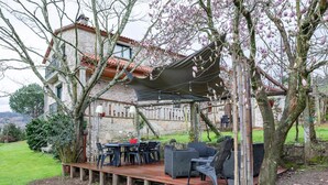 A wooden deck with outdoor furniture, a stone building with a tiled roof, and flowering trees. - Apartment 'Casa Rural Os Carballos' with Private Pool, Wi-Fi and Air Conditioning (Pontevedra)