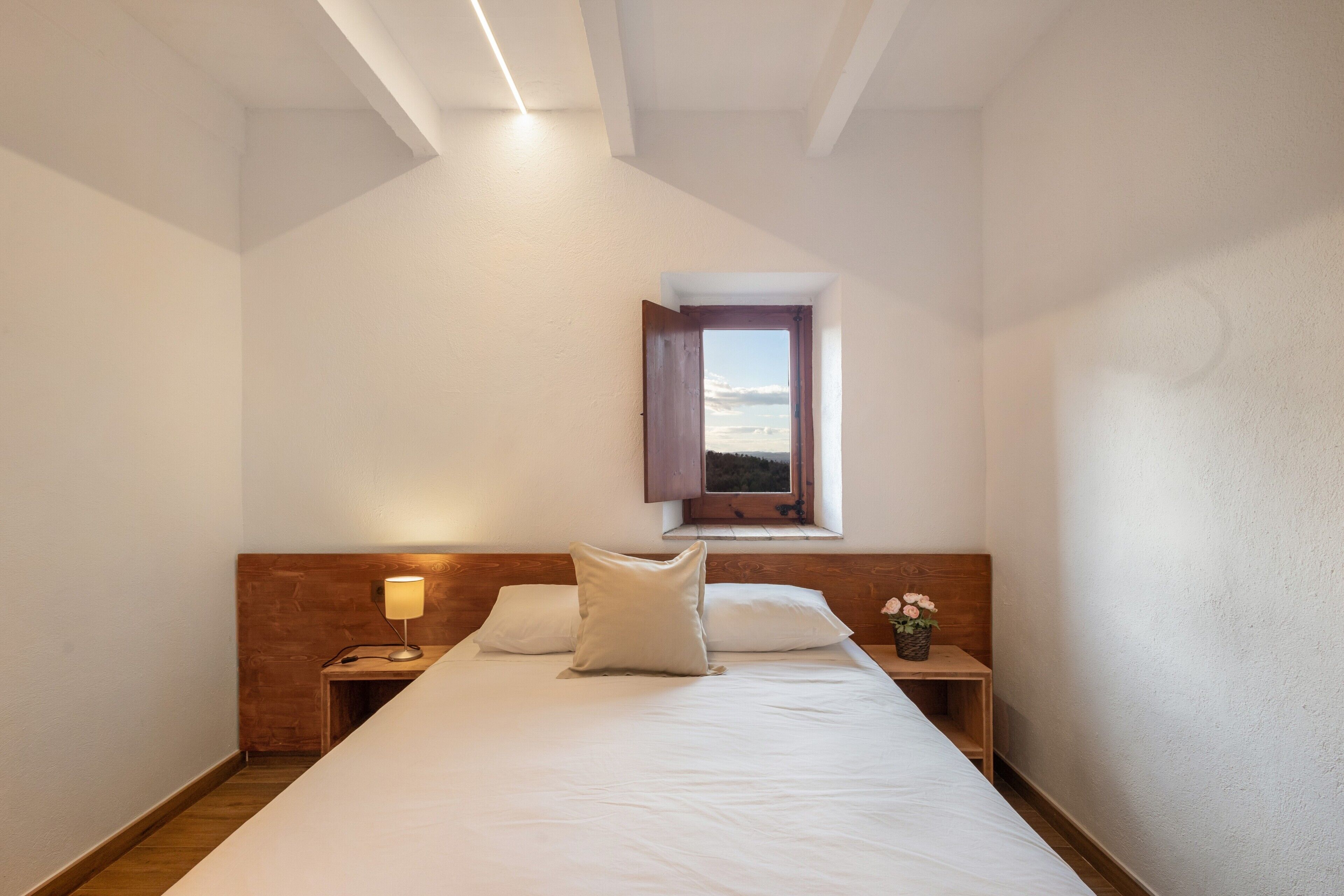 Ein Schlafzimmer mit Bett, Nachttisch, Lampe, Fenster und Blick zum Himmel.
