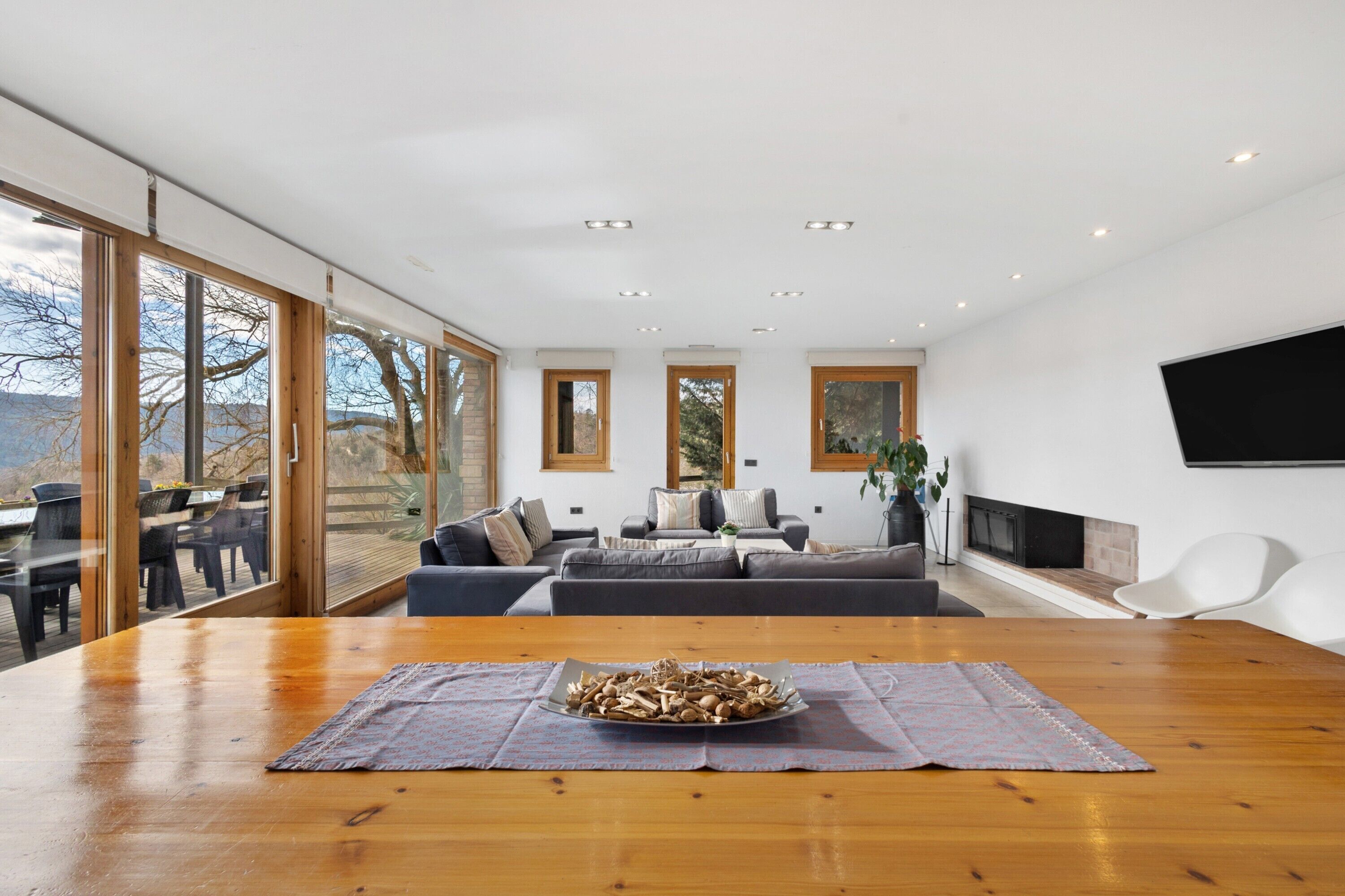 Un vaste espace de vie comprenant une grande table en bois, une cheminée et de grandes fenêtres donnant sur l’extérieur.