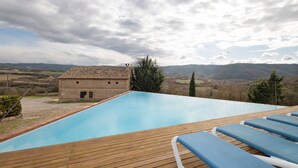 Piscina con cubierta de madera y sillones de descanso, un edificio de piedra y vistas a las colinas.
