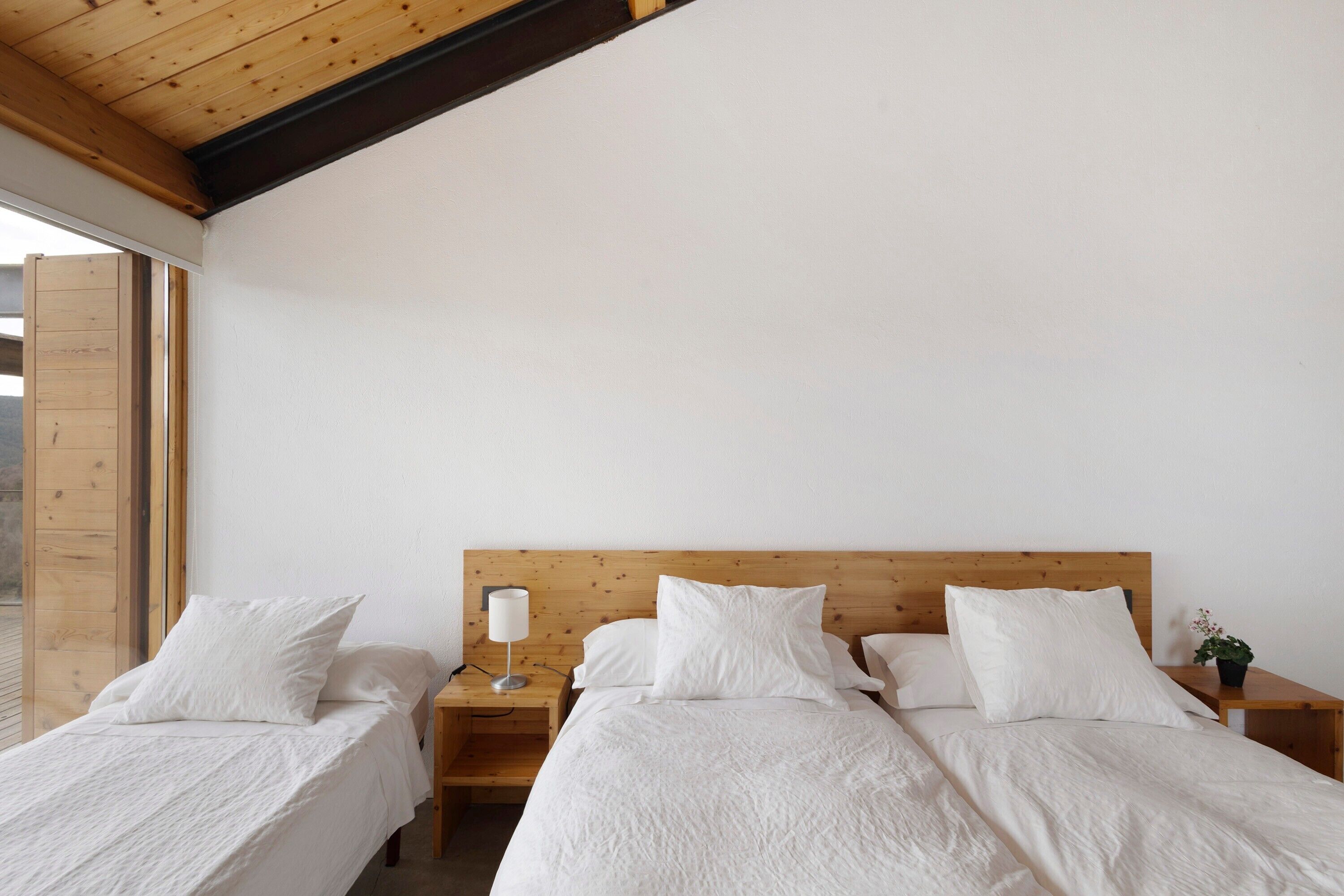 Una habitación con dos camas individuales, cabeceros de madera, una mesita de noche con lámpara y un ventanal con marco de madera.