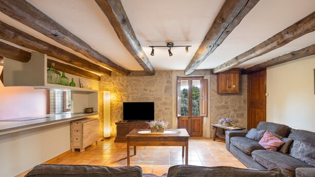 A living room with a sofa, a coffee table, and a television. There is a kitchen area with a counter and cabinets. The room has exposed wooden beams on the ceiling.
