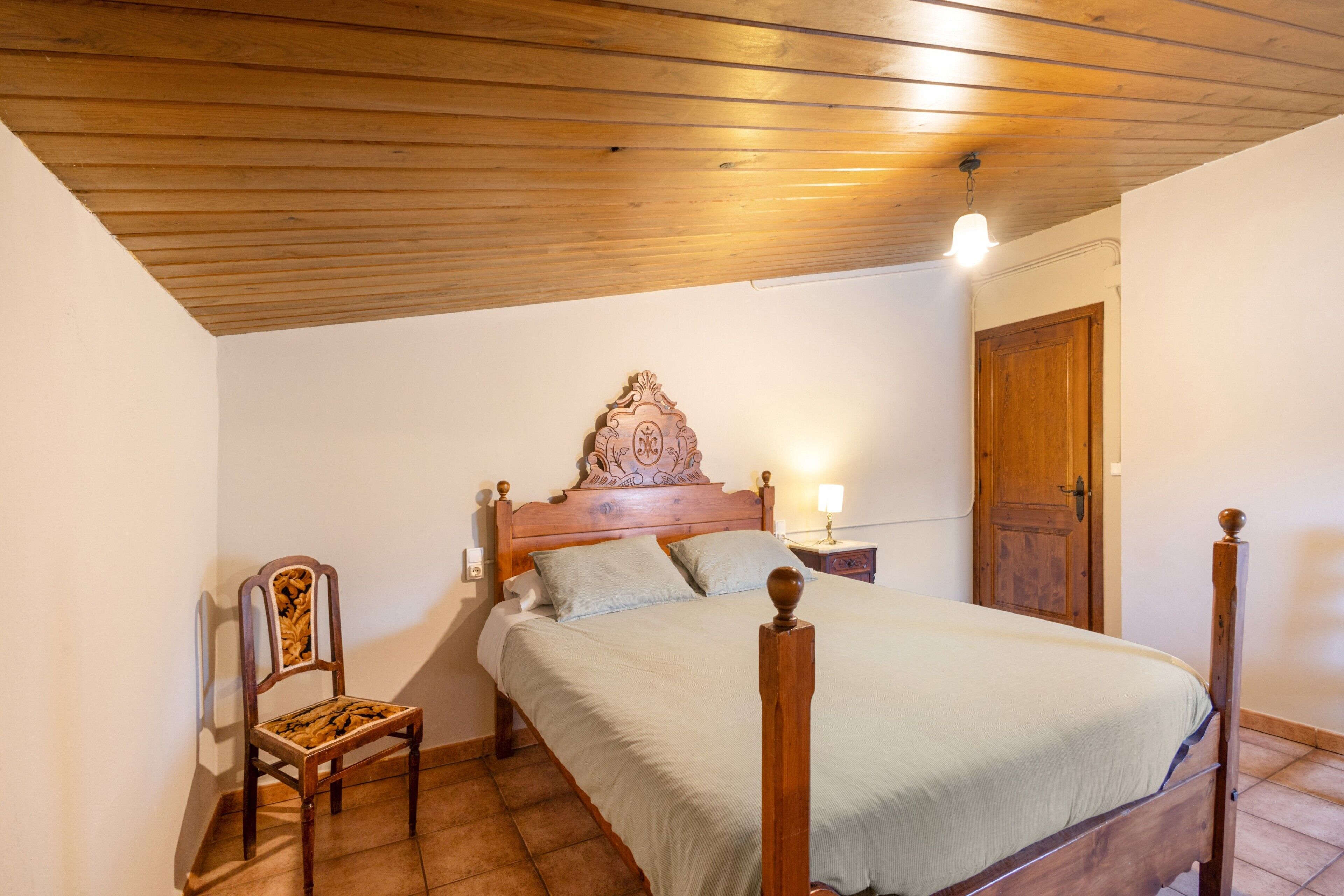 Un dormitorio con techo de madera, una cama con cabecera, una mesita de noche, una silla y una puerta.