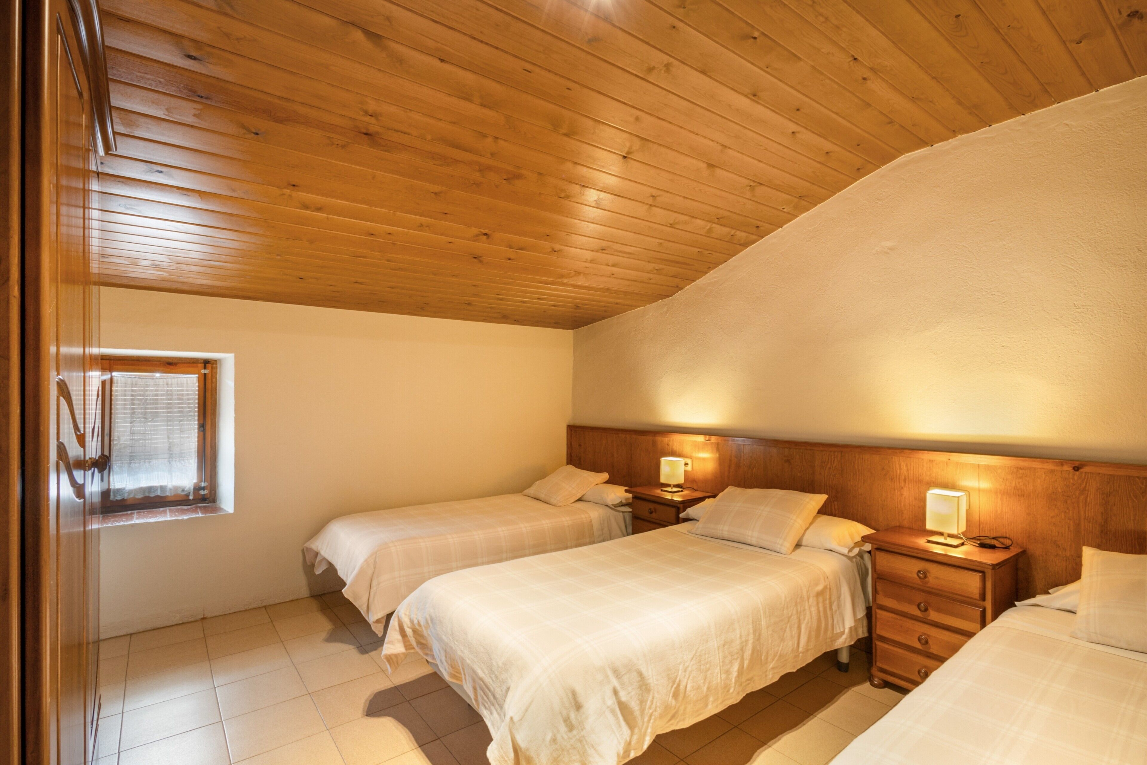 A bedroom with two beds, wooden headboard, bedside tables with lamps, a window, and a door.