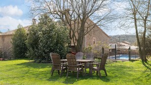 An outdoor dining area with a table and chairs, surrounded by trees and a building with a tiled roof.