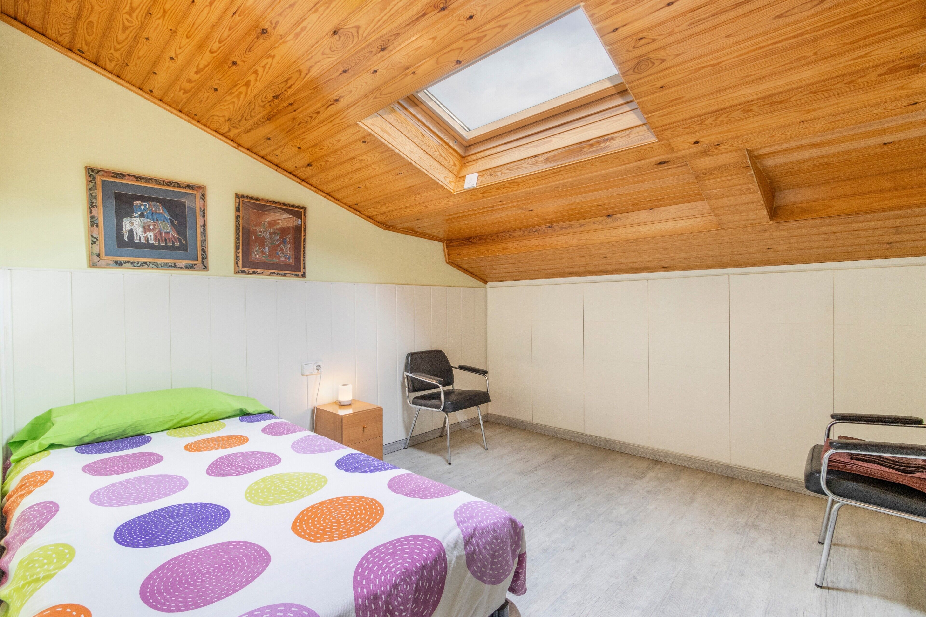 Un dormitorio con cama, una silla, una mesita de noche, tragaluz y una obra de arte en la pared.